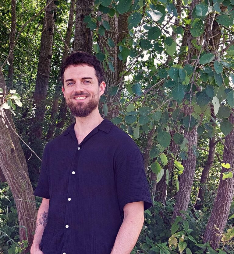 Smiling person with a black short sleeve shirt, a tattoo on the right arm. In the background there are trees and green leaves.